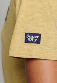 Gul kortärmad skjorta med en liten mörkblå "Super Dry"-tyglapp nära ärmslutet på en persons arm.