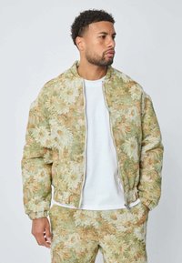 Homme portant une veste à motif floral vert et beige et un pantalon assorti par-dessus un t-shirt blanc, regardant sur le côté avec les mains dans les poches.