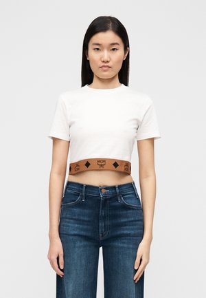 Λευκό κοντομάνικο crop top με κοντά μανίκια και καφέ ελαστική ζώνη μέσης με το λογότυπο MCM και σχέδια με μαύρα διαμάντια. Φοριέται με σκούρο μπλε τζιν.