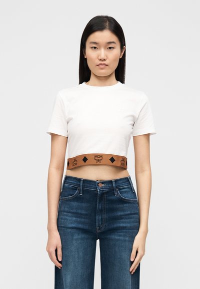 T-shirt blanc court à manches courtes avec une ceinture élastique marron ornée du logo MCM et de motifs en forme de diamant noir. Porté avec un jean bleu foncé.