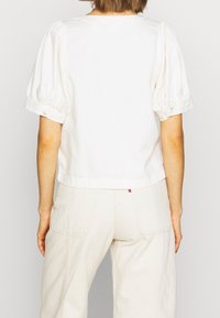 Haut blanc à manches courtes bouffantes, encolure ronde, et coupe courte. Porté avec un pantalon taille haute beige clair.