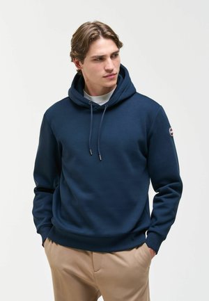 Giovane uomo con felpa blu navy con coulisse e pantaloni beige, in piedi con le mani in tasca, guarda di lato su uno sfondo semplice.