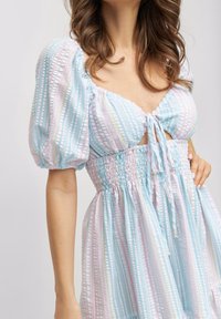 Hellblaues Kleid mit puffigen Ärmeln, gerüschtem Oberteil und vorn bindbarem Design. Es hat pastellfarbene Streifen und einen schwingenden Rock. Das Material wirkt strukturiert.