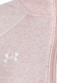 Chaqueta rosa claro con cremallera, hecha de un tejido suave y texturizado. Presenta un logo bordado en blanco y costuras visibles a lo largo de la cremallera.