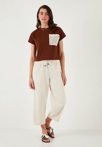 Brown cotton T-shirt with a white crocheted pocket; cream wide-leg trousers; brown sandals with decorative accents; minimalistic design.