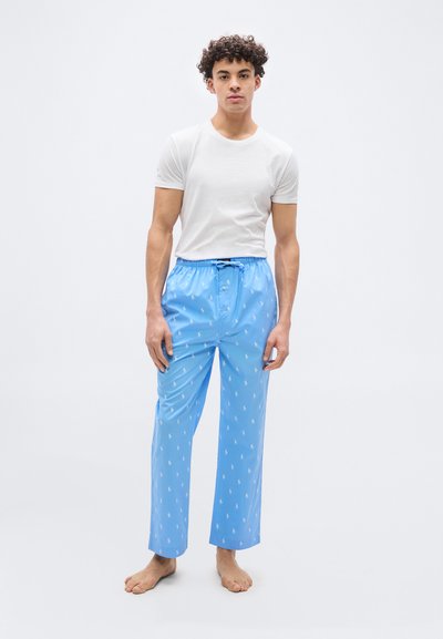 Νεαρός άνδρας όρθιος, ξυπόλητος, ντυμένος με λευκό t-shirt και ανοιχτόχρωμο μπλε παντελόνι πιτζάμα με διακριτικό λευκό μοτίβο, μπροστά σε απλό φόντο.