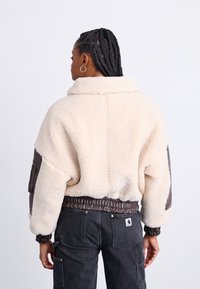 Giacca in shearling color crema di lusso con dettagli in pelle marrone, colletto alto, vita elasticizzata e vestibilità oversize; vista dorsale mostrata.