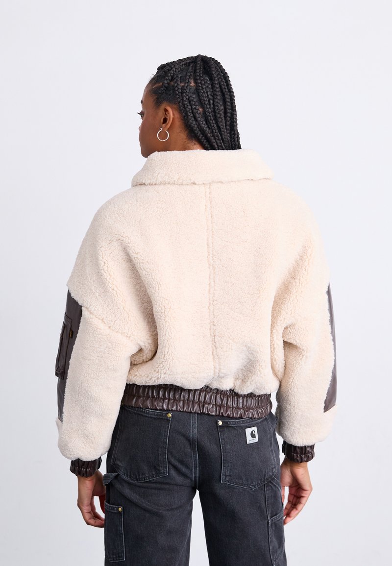 Luxuriöse, cremefarbene Lammfelljacke mit braunen Lederakzenten, hohem Kragen, elastischem Bund und oversize Passform; Rückansicht gezeigt.