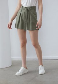 Pantaloni scurți verzi din in cu un design plisat și un brâu de legat, asortați cu adidași albi. Textură netedă și lungime până la mijlocul coapsei.