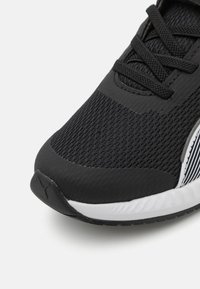 Chaussure de sport noire avec une tige en mesh, des superpositions texturées, des lacets noirs et une semelle intermédiaire blanche avec un accent rayé sur le côté.