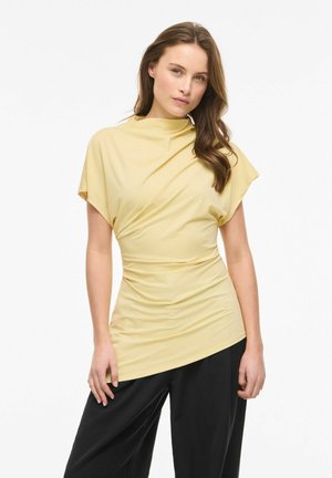 Jeune femme debout, portant un haut froncé jaune clair à manches courtes et un pantalon noir ample, sur un fond blanc.