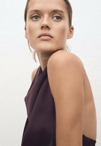 Satin purple top with a draped design, featuring a wide neckline and open shoulders, against a neutral background. Smooth texture visible.