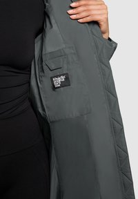 Personne tenant une veste grise matelassée ouverte montrant une poche intérieure avec une étiquette Khujotm Original For A Few.