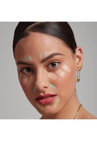 Des échantillons de cosmétiques sur le visage incluent des teintes beige clair, pêche et verte. Le modèle a la peau lisse et porte des boucles d'oreilles en forme de créoles dorées.