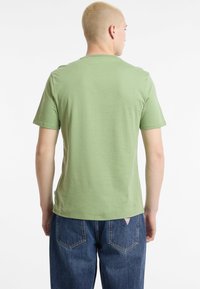 Zaļš kokvilnas t-krekls ar īsām piedurknēm, apaļu kakla izgriezumu un gludu tekstūru, valkāts kopā ar ziliem augstām jostasvietām džinsiem ar aizmugurējām kabatām.
