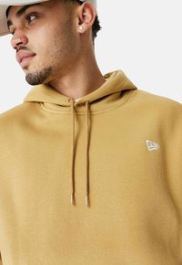 Felpa marrone con cappuccio regolabile, polsini a costine e un piccolo logo bianco sul petto. Tessuto morbido e texture.