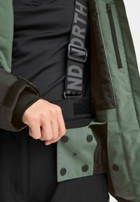 Grüne und schwarze Oberbekleidung mit verstellbaren Riemen, Druckknöpfen und einer Reißverschlusstasche. Der Stoff hat eine strukturiert-geschichtete Optik.