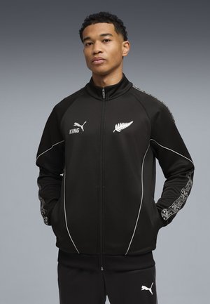 Ung mand iført sort Puma jakke med hvid bregnelogo og geometrisk mønster på ærmerne, stående med hænderne i lommerne.