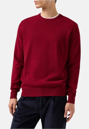 Uomo che indossa un maglione rosso semplice con scollo tondo, polsini e orlo a coste, sopra una maglietta bianca, abbinato a pantaloni scuri.