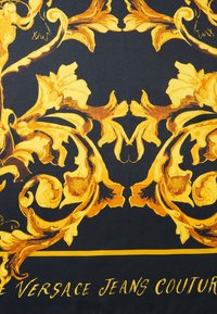 Musta kangas, jossa on monimutkainen kultainen barokkikuosis, joka sisältää kieppuvia koristeita. "Versace Jeans Couture" -teksti kultaisena alaosassa.