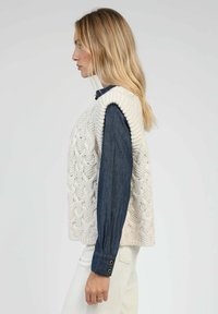 Gilet en tricot de couleur crème avec un motif de câble texturé, porté sur une chemise en denim bleu, à manches longues et avec des détails de boutons.