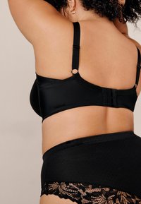 Zwart lingeriesetje met een gladde bh met verstelbare bandjes en haaksluiting, gecombineerd met hooggeknoopte slipjes versierd met kantdetails.