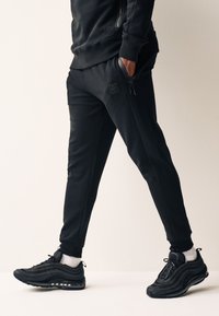 Zwart sportief trainingspak met aangesloten joggers, voorzien van ribbels bij de boorden en ritszakken, gecombineerd met zwarte sneakers en witte enkelsokken.