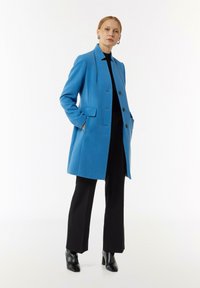 Helleblaue Jacke mit geradem Schnitt, einreihigem Design, zwei frontalen Taschen und schwarzen Knöpfen, kombiniert mit schwarzen weit geschnittenen Hosen.