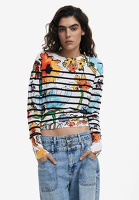 Langärmliges florales Oberteil mit leuchtenden Farben und schwarzen Streifen, aus leichtem Stoff gefertigt. Kombiniert mit hochgeschnittenen blauen Jeans.