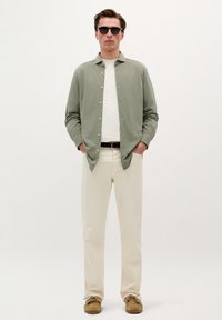 Cămașă verde deschis cu nasturi pe deasupra unei tricouri albe, asociată cu pantaloni bej și pantofi maro; ochelarii de soare completează ținuta.
