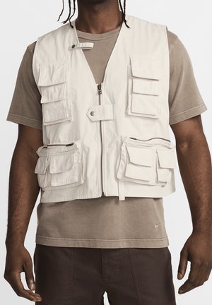 Man draagt een beige werkvest met meerdere klepzakken en een rits aan de voorkant over een bruin T-shirt en donkere broek.