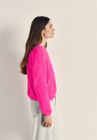 Moteris su ilgais rudos spalvos plaukais, dėvinti ryškiai rausvą megztinį ir šviesiai mėlynas džinsines kelnes, stovinti šonu priešais vienspalvį smėlio spalvos foną.