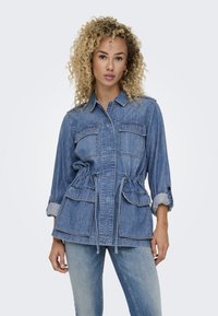 Veste en denim à manches longues, avec deux poches poitrine, taille à cordon de serrage et poignets retroussés. Présente un lavis bleu clair et une texture douce.