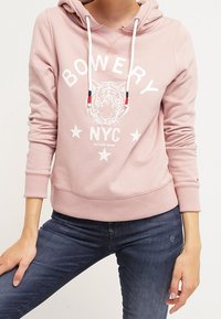 Person som bär en ljusrosa hoodie med vita dragband och texten "BOWERY NYC", tigergrafik och stjärnor, ihop med mörkblå jeans.