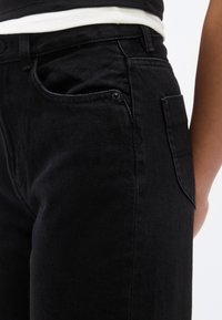Nahaufnahme einer Person, die schwarze Jeans aus Denim und ein weiß-schwarz gestreiftes Hemd trägt, mit sichtbaren Front- und Seitentaschen.