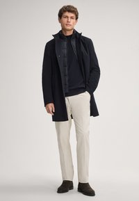 Schwarzer Überwurf mit einer taillierten Innweste, kombiniert mit einem marineblauen Pullover und cremefarbenen Hosen. Braune Wildleder-Stiefeletten runden das Outfit ab.