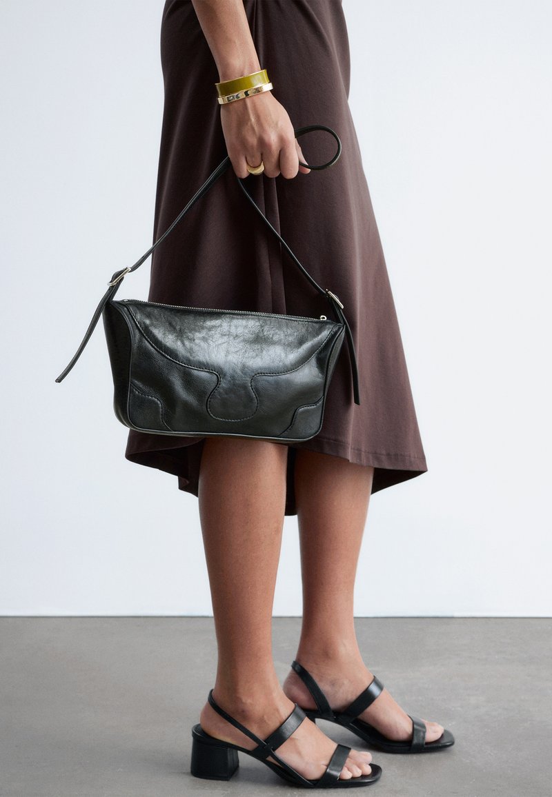 Femme portant une robe marron et des sandales à talons noires tenant un sac à main en cuir noir par sa bandoulière devant un fond uni.