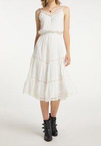 Weisses, gestuftes Midi-Kleid mit Spitzenverzierungen; leichtes Material, dünne Träger und lockere Passform. Kombiniert mit schwarzen Knöchelstiefeln.