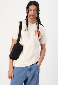 Beige bomull t-skjorte med rød grafikk på forsiden, kombinert med lyseblå denimshorts og en liten svart crossbody-veske med glidelås.