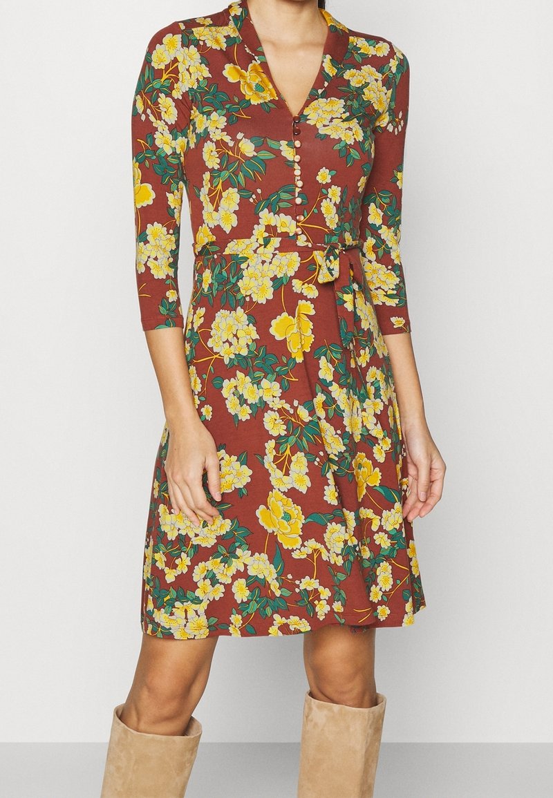 Femme portant une robe marron longueur genoux avec motif floral jaune, manches trois-quarts, boutons devant et ceinture à la taille, associée à des bottes beige.