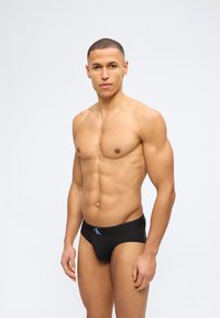 Μυώδης άνδρας στέκεται μπροστά σε απλό υπόβαθρο φορώντας μαύρα εσώρουχα Calvin Klein, κοιτάζοντας εμπρός με ουδέτερη έκφραση.