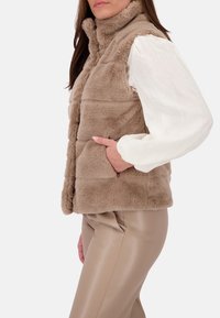 Chaleco de piel sintética en beige con un cuello alto, diseño acolchado horizontal y bolsillos laterales, combinado con una blusa crema y pantalones ajustados beige.