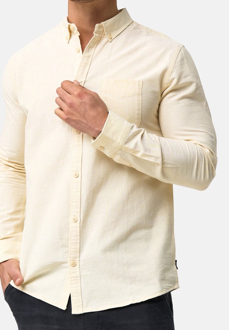 Cremefarvet langærmet skjorte i blødt stof, med en krave der kan lukkes med knapper, frontlomme og lige fald med let krumning i siderne.