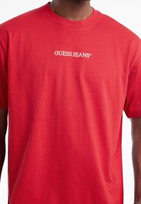 Sarkans kokvilnas T-krekls ar apkakles izgriezumu un īsām piedurknēm, ar baltu "GUESS JEANS" logo, kas izcili parādās uz krūškurvja.