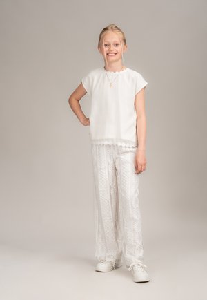 Weiße Bluse mit schmalen Ärmeln und gewelltem Saum, kombiniert mit weißen gemusterten weiten Hosen. Weiße Sneaker runden das Outfit ab.