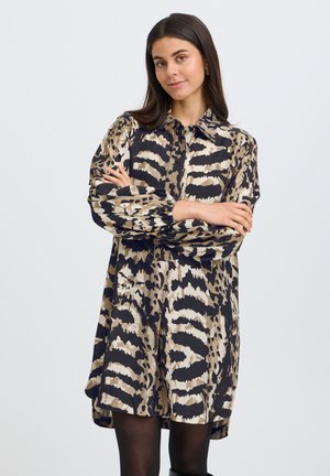 Femme aux longs cheveux foncés portant une robe à imprimé animal noir, beige et fauve à manches longues, bras croisés, sur un fond uni clair.