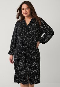 Czarna sukienka z guzikami na plecach, zdobiona białymi kropkami, z długimi rękawami, warstwowym talią i średniej długości hemline'm z rozcięciami z przodu.