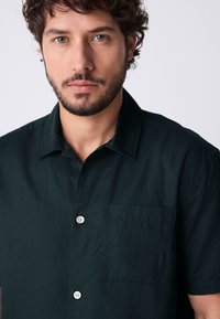 Camisa de manga corta verde oscuro hecha de tela texturizada, que cuenta con un cierre de botón en la parte delantera y un bolsillo en el pecho izquierdo con un solo botón.