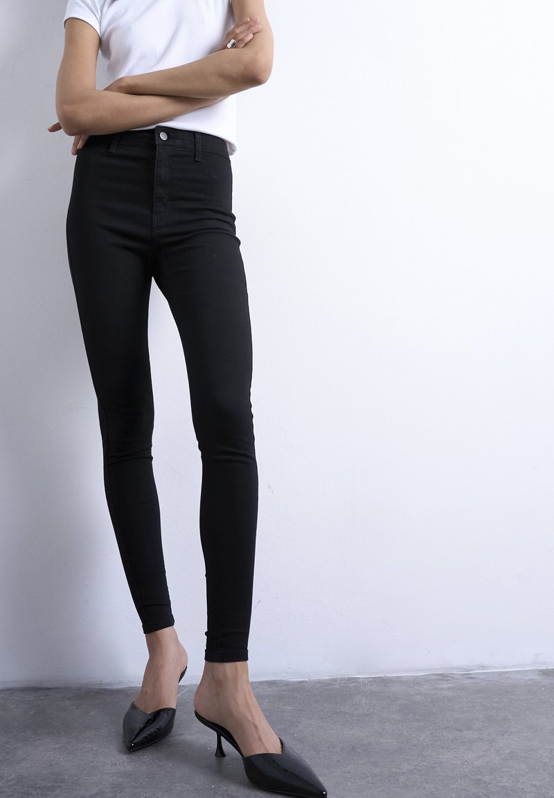 Personne portant un pantalon noir ajusté, une chemise blanche à manches courtes et des mules à talons noires à bout pointu, debout les bras croisés.