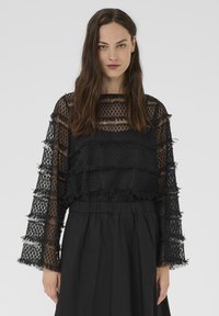 Sieviete ar gariem brūniem matiem, vilkusi melnu caursp�īdīgu mežģīņu topu ar frizūru detaļām un augsta vidukļa melnām svārkiem uz vienkārša fona.